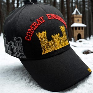 May include: Black baseball cap with the words "COMBAT ENGINEER" embroidered in red. The cap features a gold and black castle design on the front and side, with a black brim. The cap is sitting on a snowy surface.