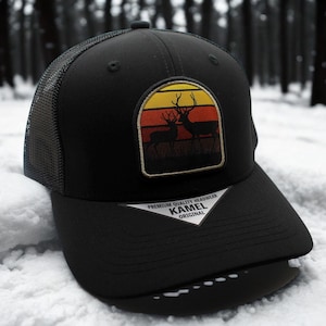 May include: Black trucker hat with a mesh back and a curved brim. The front features a patch with a sunset design and two deer silhouettes. The hat has the text "KAMEL ORIGINAL" on the tag.