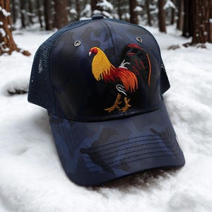 May include: A navy blue camouflage baseball cap with a mesh back. The cap features an embroidered rooster in yellow, red, and orange. The hat is set against a snowy, wooded background.