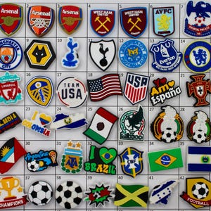 May include: A collection of 47 colorful shoe charms featuring various sports team logos, flags, and soccer balls. The charms are made of rubber and have a variety of designs, including the Arsenal, West Ham, Liverpool, and USA logos, as well as flags from countries like Colombia, Mexico, and Brazil.