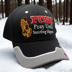 May include: Black baseball cap with the embroidered text "PUSH Pray Until Something Happens." The cap features a gray bill and a design of praying hands with a cross. The background is a snowy forest.