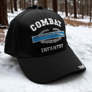 Könnte beinhalten: Schwarze Baseballkappe mit dem Wort "COMBAT" in weißer Stickerei über einem blauen und silbernen Emblem und dem Wort "INFANTRY" darunter. Die Kappe hat einen gebogenen Schirm und ein kleines gesticktes Detail an der Seite.