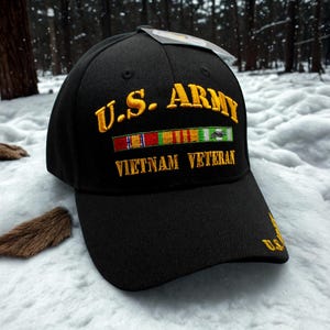 Może przedstawiać: Czarna czapka bejsbolowa ze złotym haftowanym napisem "U.S. ARMY" i "VIETNAM VETERAN". Czapka posiada rząd kolorowych haftowanych wstążek. Daszek również ma haftowany tekst. Czapka jest na tle śnieżnego tła.