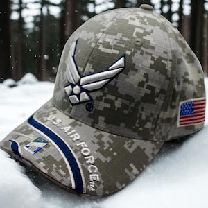 May include: A camouflage baseball cap featuring the U.S. Air Force logo and the American flag. The cap has a digital camo pattern in shades of gray, green, and tan. The words "U.S. AIR FORCE" are embroidered on the brim.