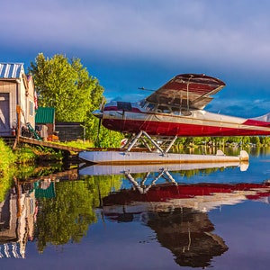 May include: A red and white seaplane with floats is parked on a lake. The plane has the letters "MT" on the tail. A small wooden building is on the shore.