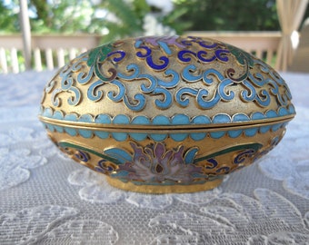 May include: A gold oval-shaped trinket box with a blue, green, and purple floral design. The box is decorated with intricate swirls and dots.