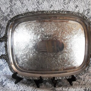 May include: A silver-colored, rectangular serving tray with ornate floral and scrollwork detailing. The tray has two curved handles and a decorative border. The tray is displayed on a black stand against a white lace background.