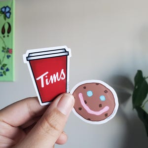 Puede incluir: Dos pegatinas, una de una taza de café roja con la palabra "Tims" y la otra de una galleta de chocolate con chispas marrón con una carita sonriente rosa.