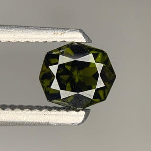 Könnte beinhalten: Ein facettierter olivgrüner Edelstein in sechseckiger Form, der von einer silberfarbenen Metallpinzette gehalten wird. Der Stein weist in der Mitte ein ausgeprägtes sternförmiges Muster auf, das durch die Anordnung der Facetten entsteht.