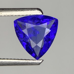 May include: A vibrant, deep blue gemstone cut into a triangular shape with multiple facets that reflect light. The stone is held securely between the two metal prongs of a pair of tweezers, showcasing its intense color and geometric clarity.