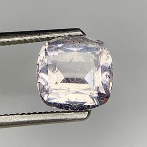 Puede incluir: Una piedra preciosa de corte cojín con un tono lavanda pálido y superficie facetada, sostenida por unas pinzas de metal. La piedra muestra claridad interna con facetas visibles que reflejan la luz, creando un patrón geométrico en su cuerpo.