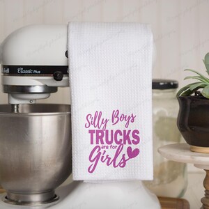 Op de afbeelding: Witte keukendoek met roze tekst die "Silly Boys Trucks are for Girls" met een roze hartje zegt.