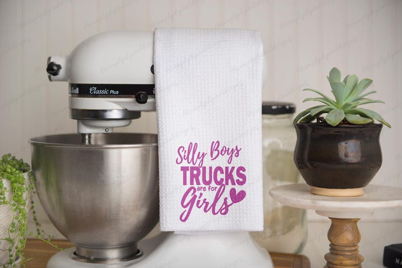 Op de afbeelding: Witte keukendoek met roze tekst die "Silly Boys Trucks are for Girls" met een roze hartje zegt.