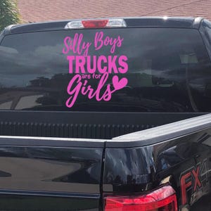 Op de afbeelding: Roze vinyl sticker met de tekst "Silly Boys Trucks are for Girls" met een roze hart. De sticker zit op de achterruit van een zwarte pick-up truck.