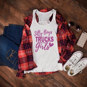 Op de afbeelding: Een wit tanktop met een paarse afbeelding waarop staat "Silly Boys Trucks are for Girls" met een roze hart. Het tanktop ligt op een houten oppervlak met een geruit overhemd, een blauwe spijkerbroek, een zonnebril en een telefoon.