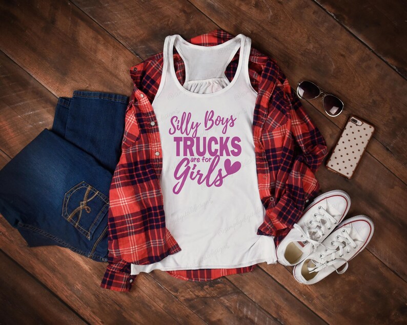 Op de afbeelding: Een wit tanktop met een paarse afbeelding waarop staat "Silly Boys Trucks are for Girls" met een roze hart. Het tanktop ligt op een houten oppervlak met een geruit overhemd, een blauwe spijkerbroek, een zonnebril en een telefoon.