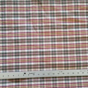 May include: A white fabric with a plaid pattern in shades of green, red, and beige. The fabric is measured with a ruler in inches.