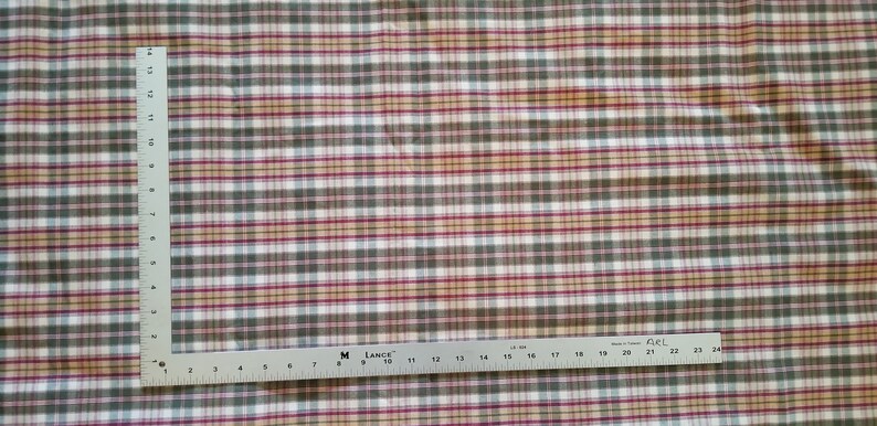 May include: A white fabric with a plaid pattern in shades of green, red, and beige. The fabric is measured with a ruler in inches.