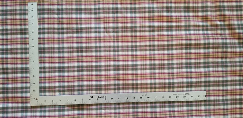 May include: A close-up of a white fabric with a plaid pattern in shades of red, green, and beige. The fabric is measured with a ruler, showing a length of 61 cm.
