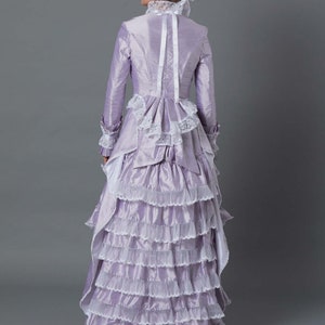 May include: A lavender gown with a white lace trim and a ruffled skirt. The gown has a fitted bodice with a high neckline and long sleeves. The gown is worn by a woman with a hat and gloves.