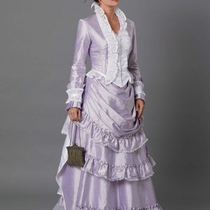 May include: A woman in a lavender dress with a white lace bodice and a ruffled skirt. She is wearing a small hat with purple flowers and a small purse.
