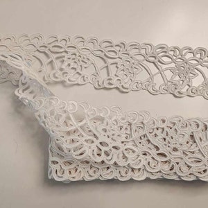 May include: Two pieces of white lace trim with a delicate floral pattern. The lace is folded and arranged on a white surface.