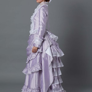 May include: A woman in a lavender dress with a white lace trim and a matching hat. The dress has a bustle in the back and a long train.