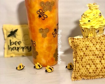 May include: A yellow and orange tumbler with a honeycomb pattern and bee designs. The tumbler has a black lid and a clear straw. There are also small bee figurines and a yellow cupcake with a bee on top. The text "bee happy" is on a yellow pillow.