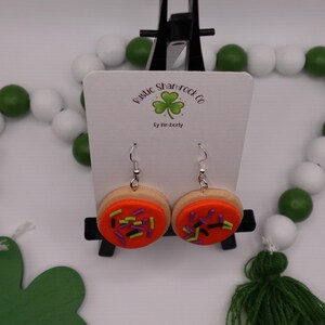 May include: A pair of handmade cookie earrings with orange frosting and colorful sprinkles. The earrings are displayed on a white card with the text "Rustic Shamrock Co" and a shamrock logo. The background includes green and white beads and a green tassel.