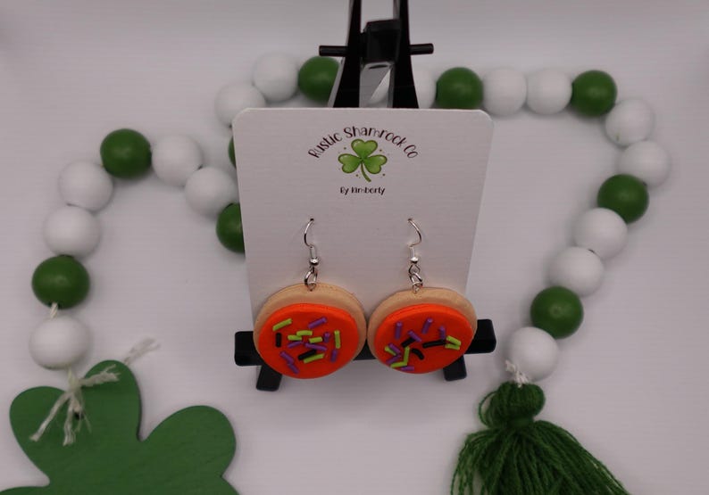 May include: A pair of handmade cookie earrings with orange frosting and colorful sprinkles. The earrings are displayed on a white card with the text "Rustic Shamrock Co" and a shamrock logo. The background includes green and white beads and a green tassel.