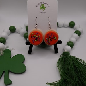 May include: A pair of dangle earrings shaped like frosted sugar cookies with orange icing and colorful sprinkles. The earrings are displayed on a white card with the text "Rustic Shamrock Co" and a shamrock logo. A green and white bead garland is in the background.