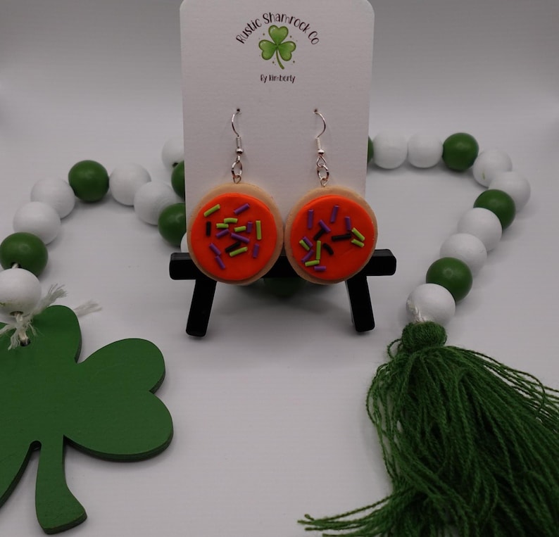 May include: A pair of dangle earrings shaped like frosted sugar cookies with orange icing and colorful sprinkles. The earrings are displayed on a white card with the text "Rustic Shamrock Co" and a shamrock logo. A green and white bead garland is in the background.