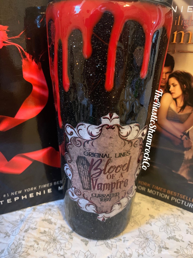Pu&ograve; includere: Un bicchiere nero glitterato con gocce rosse sulla parte superiore. Il bicchiere presenta un'etichetta che recita "Original Lines Blood of a Vampire Currated 1897". The Rustic Shamrock Co.