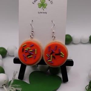 May include: A pair of dangle earrings shaped like sugar cookies. Each earring features a round, tan cookie base topped with bright orange frosting and colorful sprinkles. The earrings hang from silver-colored hooks.