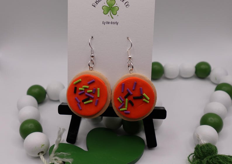 May include: A pair of dangle earrings shaped like sugar cookies. Each earring features a round, tan cookie base topped with bright orange frosting and colorful sprinkles. The earrings hang from silver-colored hooks.