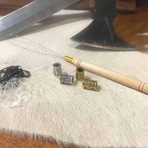 Könnte beinhalten: Haaraccessoires, darunter schwarze und transparente Gummibänder, dekorative Metallperlen und ein hölzernes Haarperlenwerkzeug. Ein großes Messer und eine Axt befinden sich im Hintergrund.