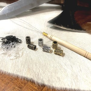 May include: A collection of hair accessories, including metal beads with engraved designs, black and clear elastics, and a wooden tool with a needle. An axe and a knife are in the background. The items are on a white fur-like surface.