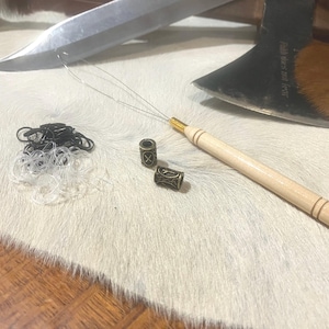 May include: A set of hair beads, a wooden tool with a metal needle, and a pile of clear hair ties on a white fur surface. The tool is used to thread hair beads onto hair.