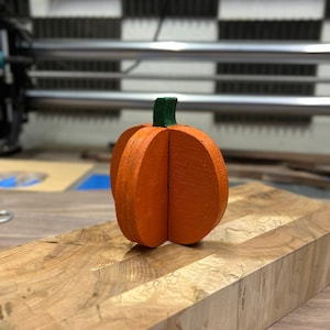 May include: A wooden pumpkin painted orange with a green stem. The pumpkin is sitting on a wooden butcher block. The pumpkin is a decorative item, perfect for fall or Halloween.