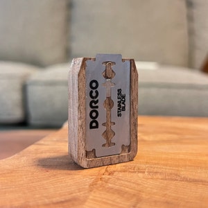 May include: A wooden box with a silver razor blade inside. The blade is labeled "DORCO STAINLESS BLADE".