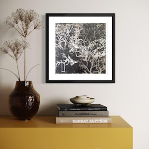 May include: A framed black and white abstract art print featuring a tree branch design. The artwork is displayed above a yellow table with a brown vase and dried flowers. Books and a small bowl are also on the table.