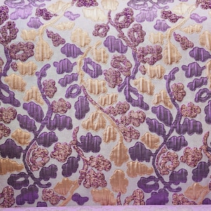 Puede incluir: Primer plano de una tela con un estampado floral. El diseño presenta flores y hojas bordadas en morado, dorado y beige claro sobre un fondo blanco. La tela tiene una apariencia texturizada.