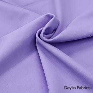 Könnte beinhalten: Nahaufnahme eines weichen, glatten, lavendelfarbenen Stoffes. Der Stoff ist gefaltet und drapiert, wodurch seine Textur zur Geltung kommt. Der Text "Daylin Fabrics" ist in der rechten unteren Ecke sichtbar.