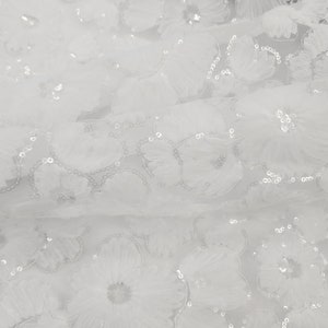 May include: White floral fabric with three-dimensional flower appliques and scattered sequins. The delicate flowers are layered, creating a textured effect. This fabric is suitable for bridal wear, evening gowns, or decorative projects.