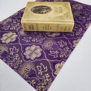 May include: A purple fabric book cover with a gold pattern of crosses, doves, and chalices. The cover is decorated with a gold border and has a gold inscription that reads "HOLY BIBLE".