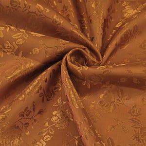 Op de afbeelding: Close-up van een gevouwen, goudbruine stof met een bloemmotief. De stof heeft een satijnglans en een gedetailleerd reliëfdesign van rozen en bladeren. De plooien creëren schaduwen die de textuur en diepte van het materiaal benadrukken.