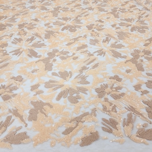 May include: Beige fabric with a floral pattern and sequins. The fabric has a delicate, sheer texture and is perfect for creating elegant garments or accessories.