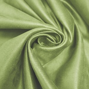 Puede incluir: Primer plano de una tela verde claro doblada, con una textura suave y aterciopelada. La tela está drapeada en un patrón arremolinado, creando sombras y reflejos que enfatizan su superficie lisa. El color es un verde apagado y natural.