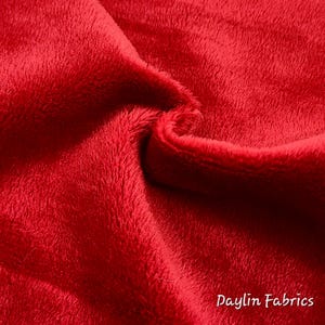 May include: Close-up of a soft, red fabric with a fuzzy texture. The fabric is folded in half, showing the inside and outside of the material. The text "Daylin Fabrics" is visible in the bottom right corner.