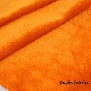 May include: Close-up of a bright orange fuzzy fabric. The fabric is soft and plush, with a subtle texture. The fabric is folded in half, showing the texture of the fabric.